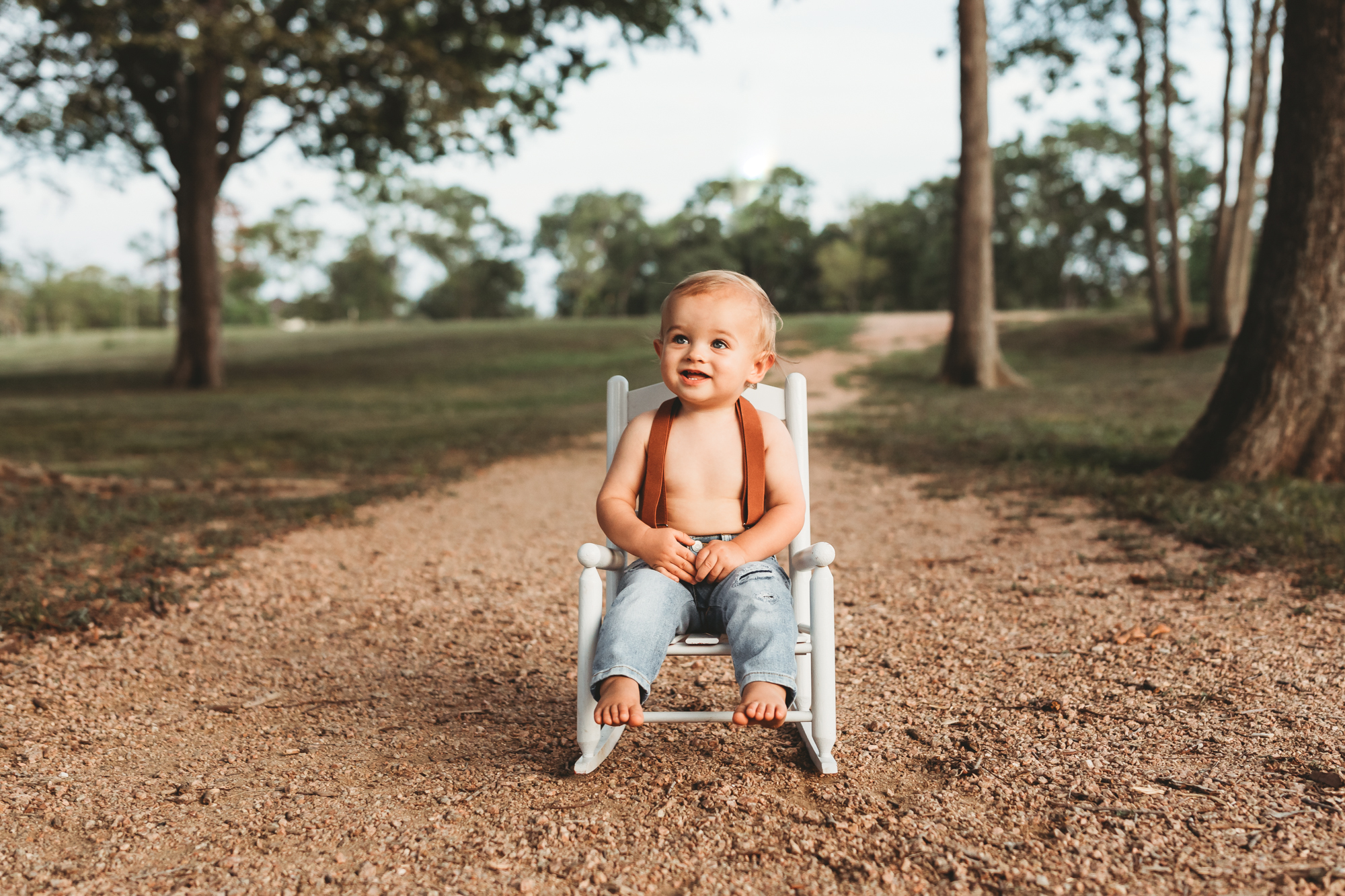 Toddler boy sitting in white rocker chair with jeans and suspenders at a park in Pearland with Houston Family Photographer.