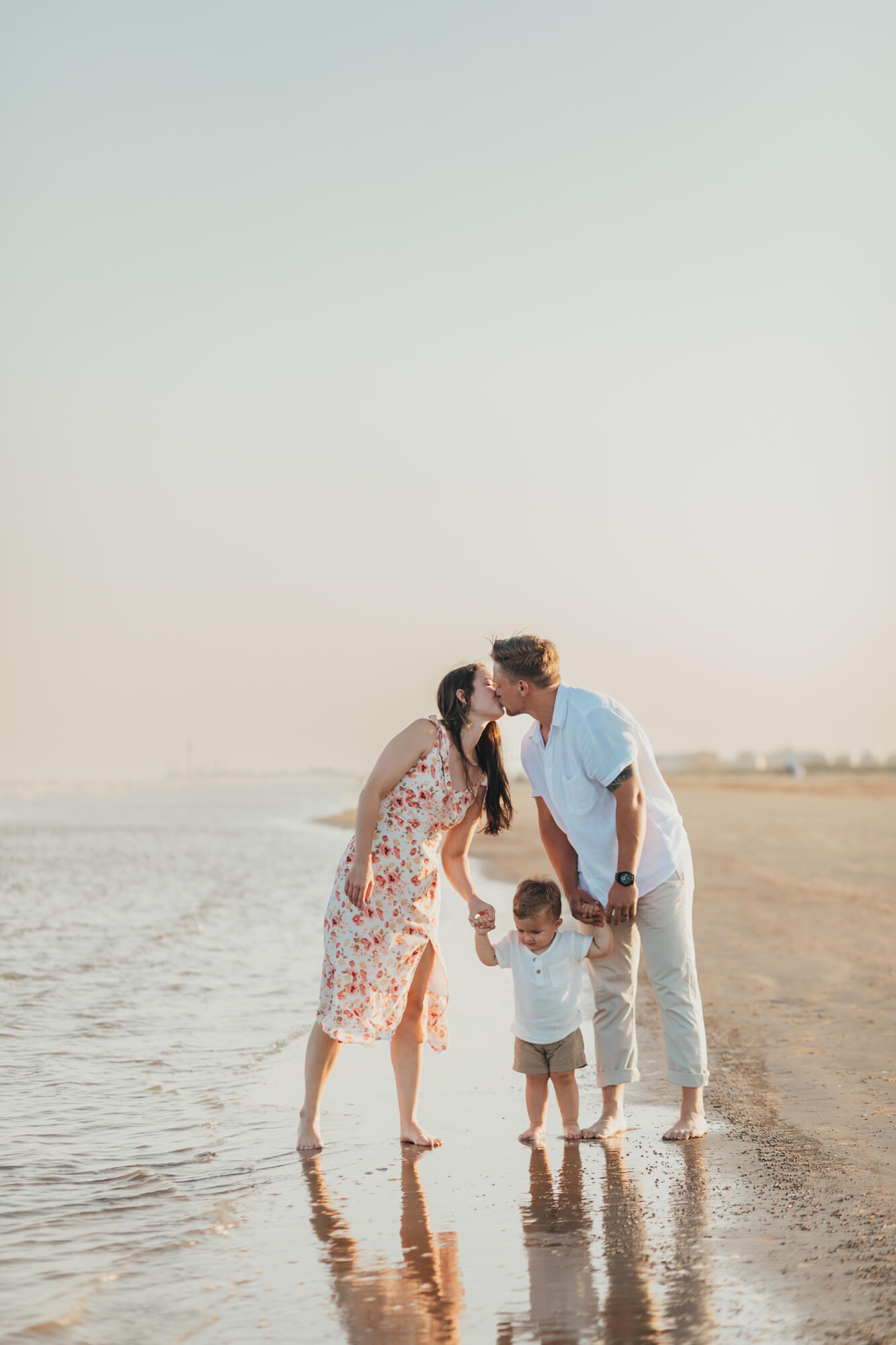 Houston restaurants with playgrounds are a win for this family, walking along the beach with their toddler in tow. Mom and dad kissing over baby boy while on the beach.