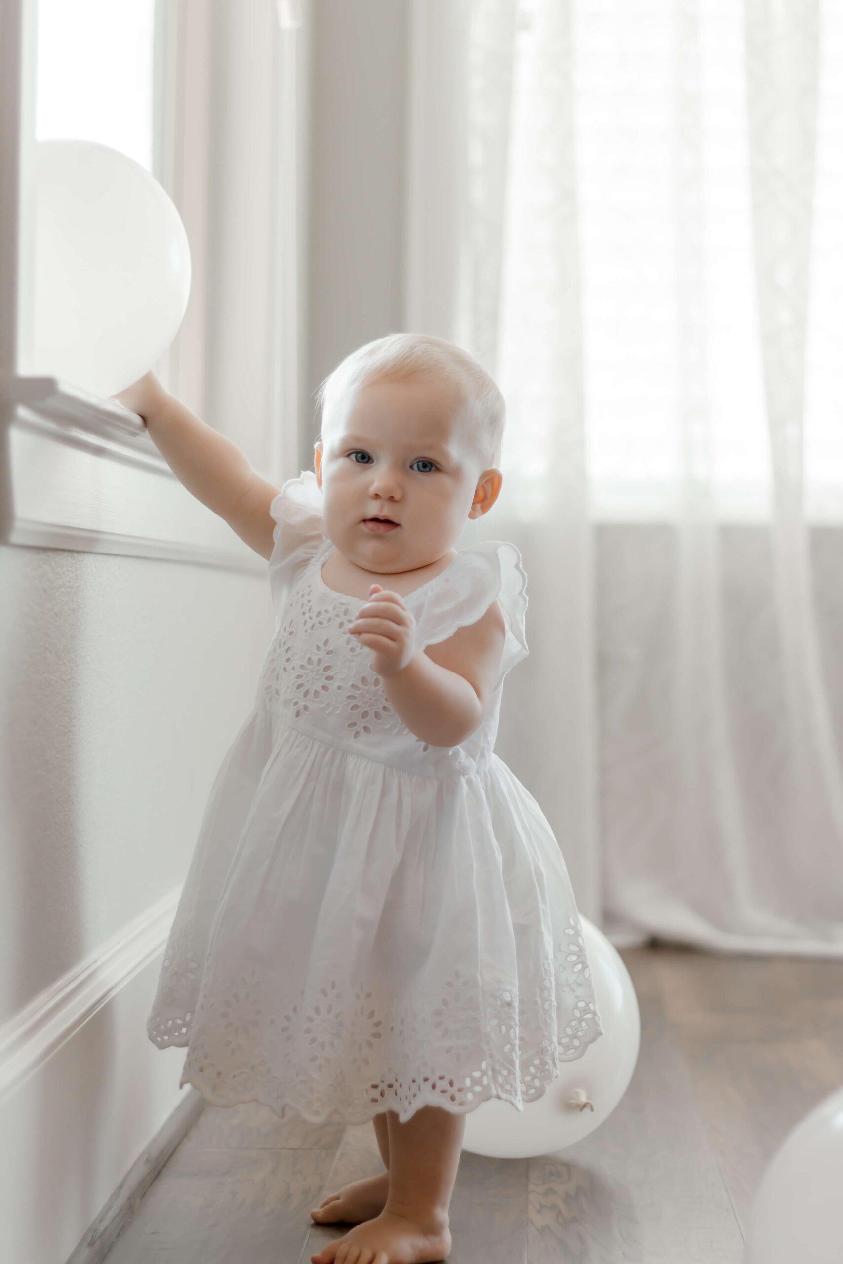 Girl playing with balloon, wearing white dress for first birthday.
