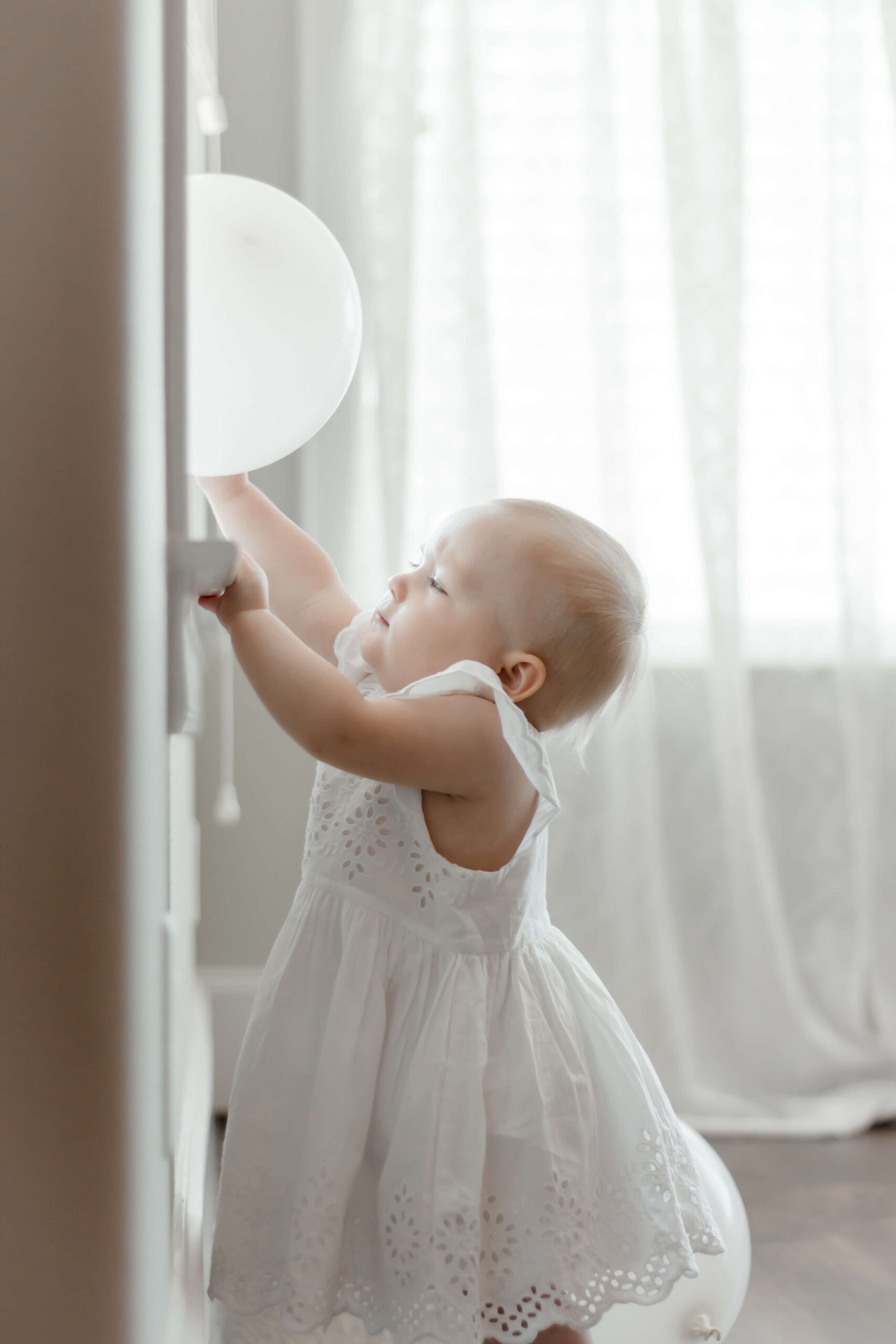 Postpartum therapy Houston is a resource that moms really need to know about. Baby girl celebrates her first birthday with a cake smash at home, wearing a white dress and playing with a white balloon in the windowsill.