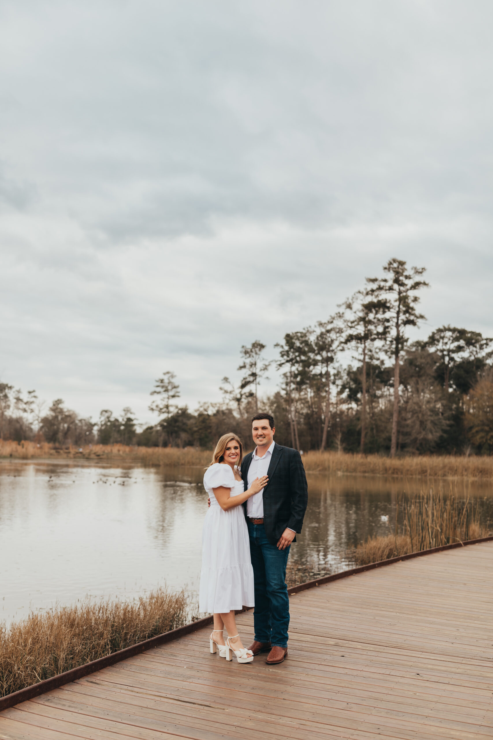 Couple hold onto each other while standing on a dock at the Brazos Bend Conservatory, with the lake in the background.